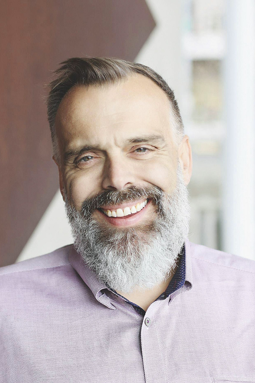 Image of a smiling, light-skinned person with short gray hair and a beard wearing a lavender button down shirt