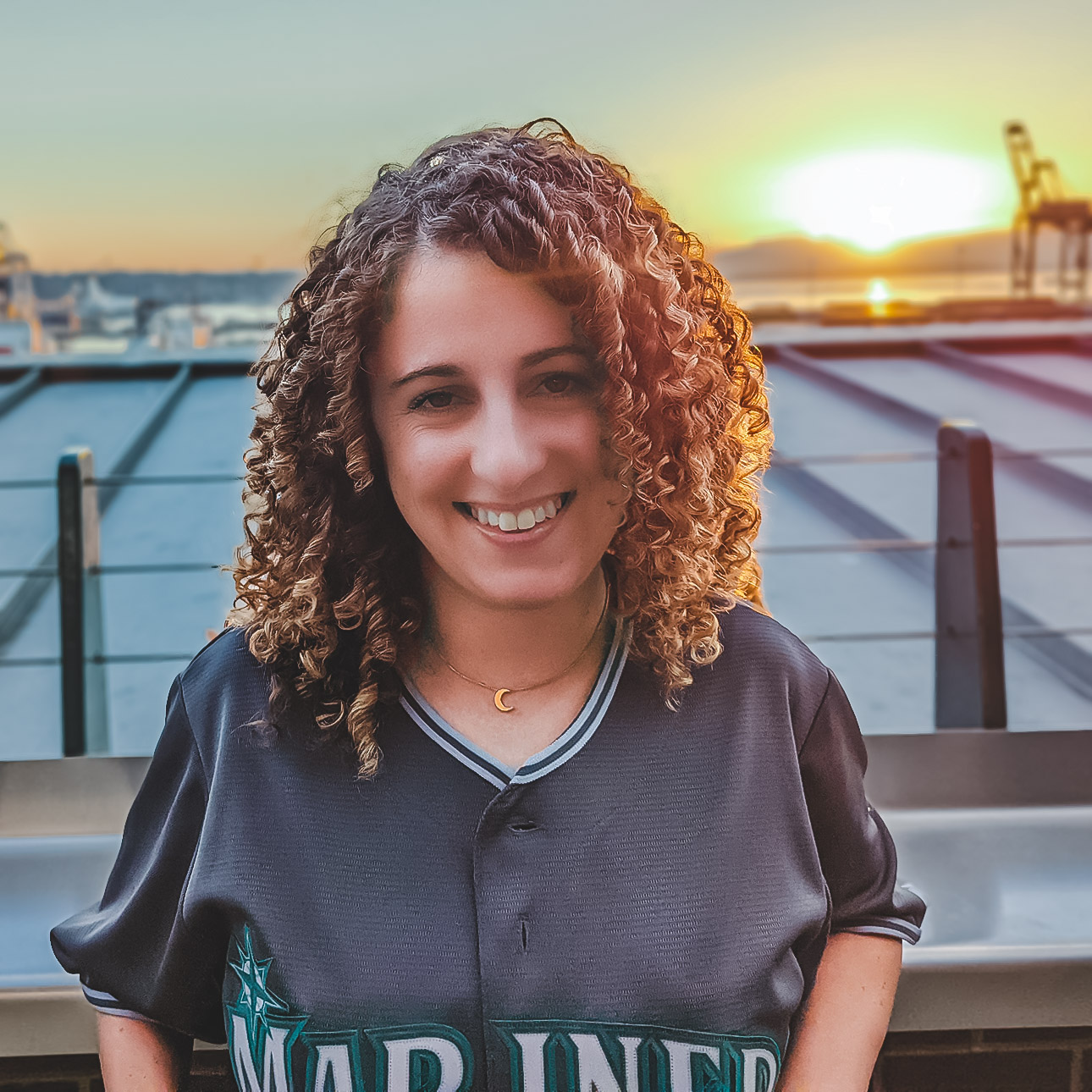 Smiling person with long, curly, brown hair wearing a Mariner jersey posing before a sunset