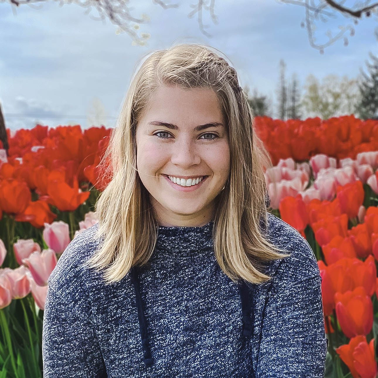 A smiling person with long blond hair wearing a sweater in a field of tulips