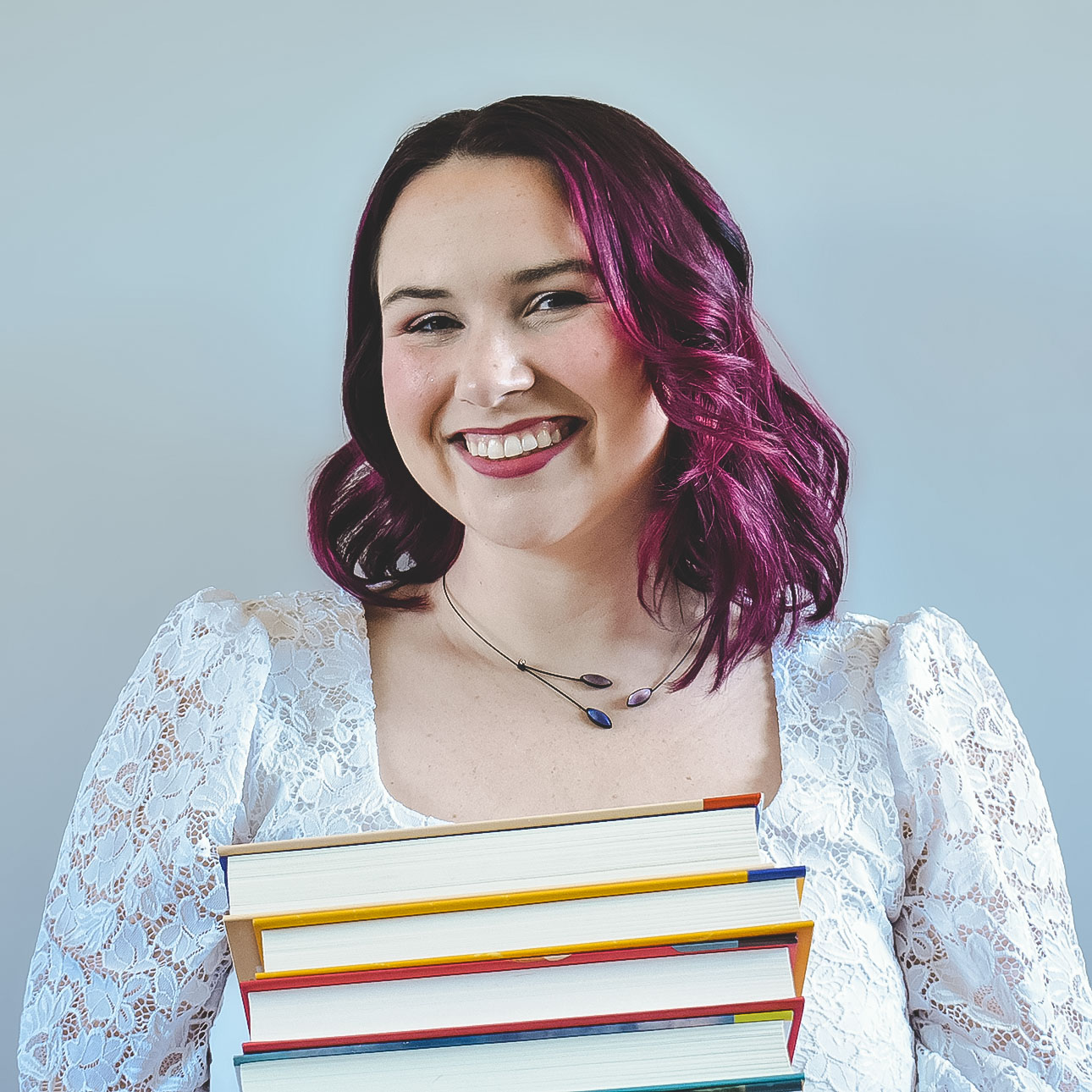 A smiling person with magenta hair wearing a white dress holding a stack of books