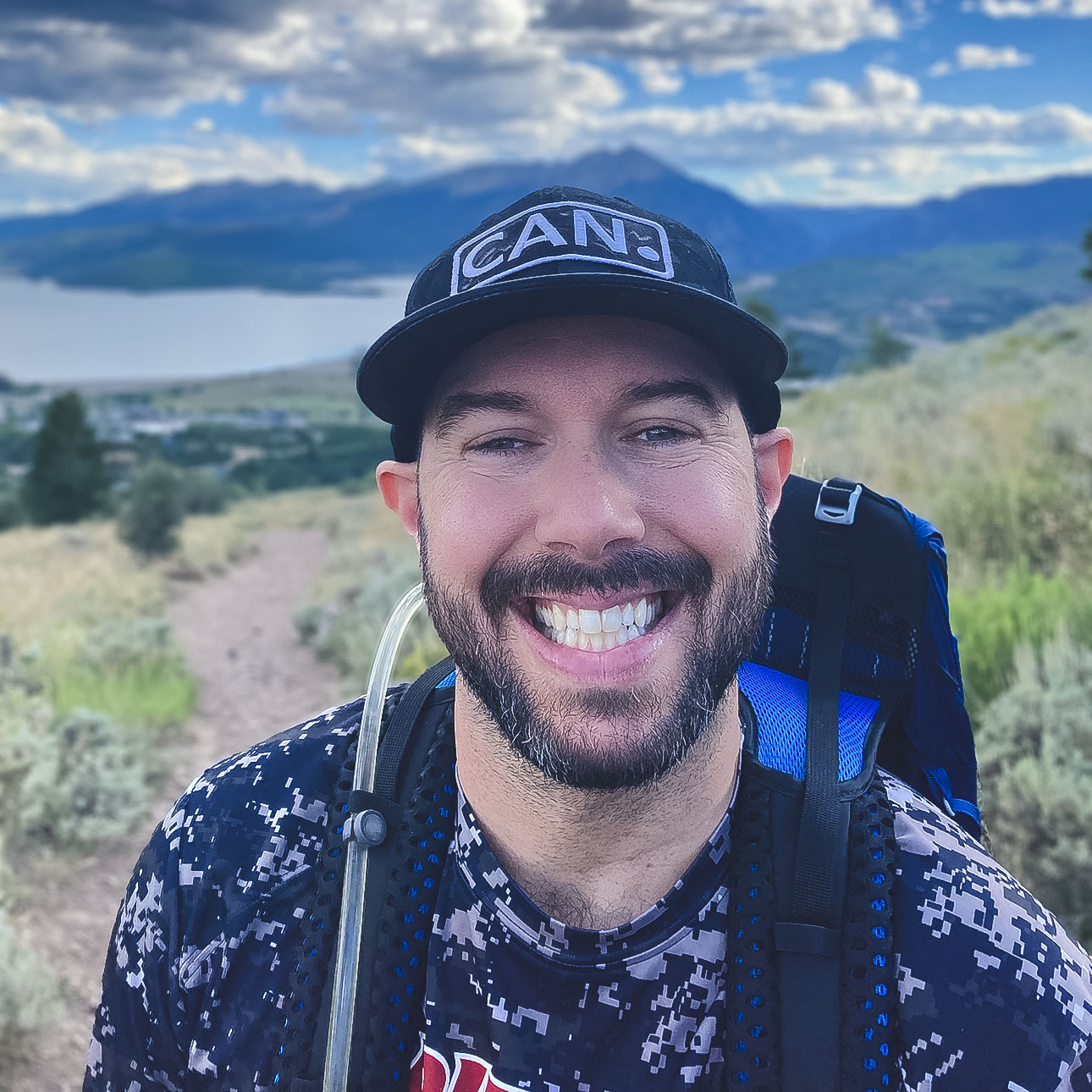 A smiling, bearded person wearing a hat and hiking pack posing before water and mountains