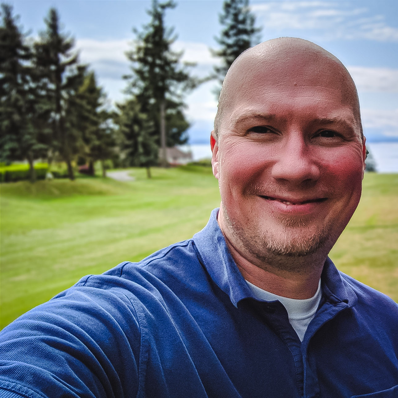 A smiling bald person wearing a blue shirt posing in a gold course