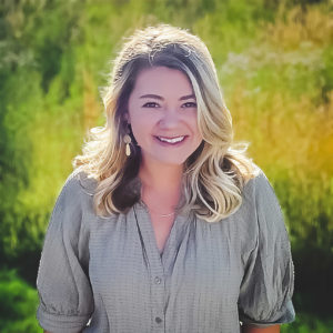 Smiling person with long, dark blond hair wearing a brown top posing for a photo before a green field