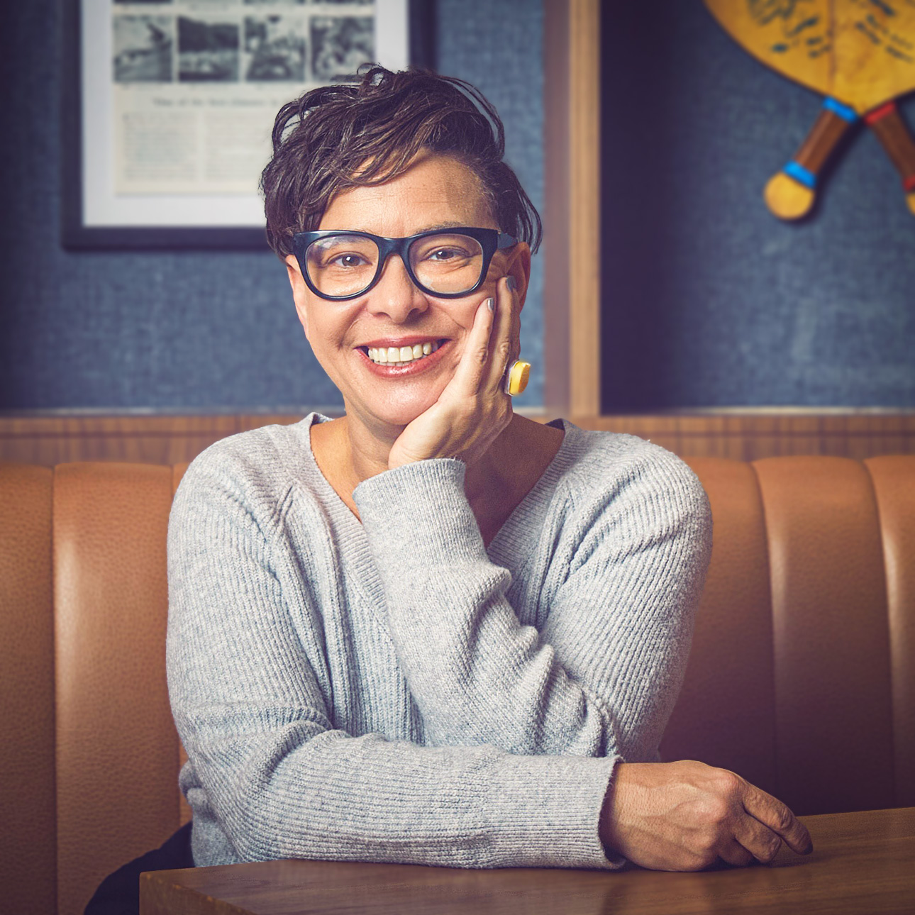 A smiling person with short dark hair wearing blue glasses posing in a dinette