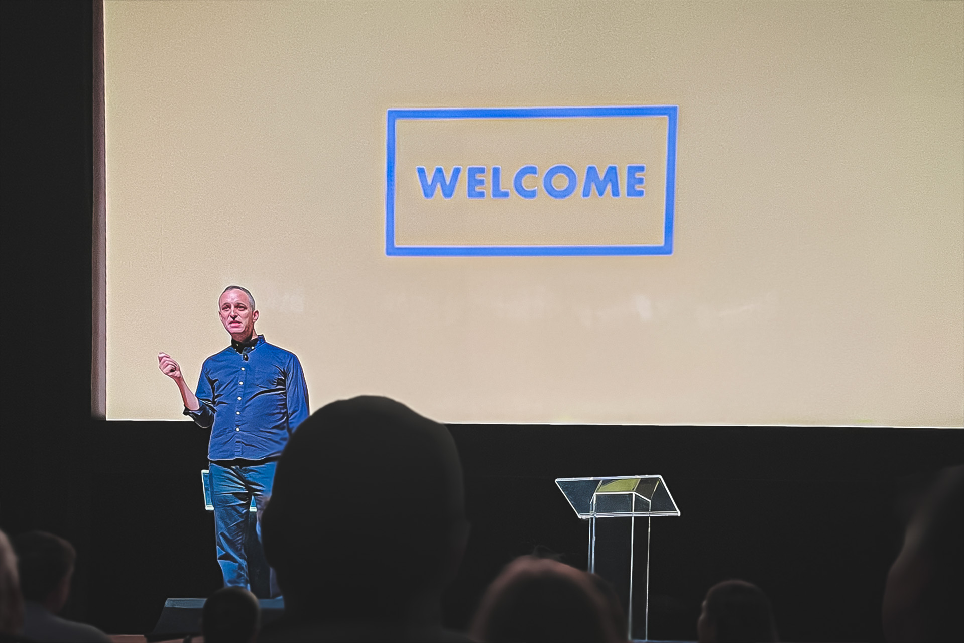 a person in a blue shirt stands before a slide presentation with the work welcome